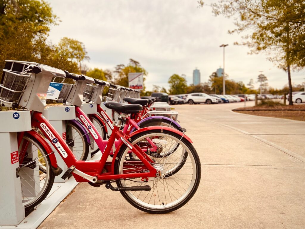 Row of red Houston BCycle bikes docked at a station with a parking lot and trees in the background.
