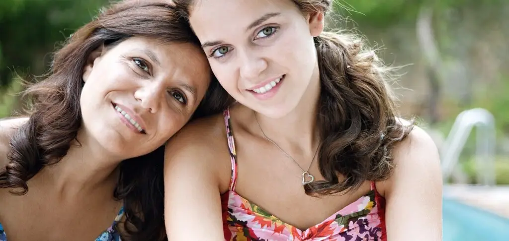 Smiling mother and teenage daughter leaning close together outdoors near a pool.