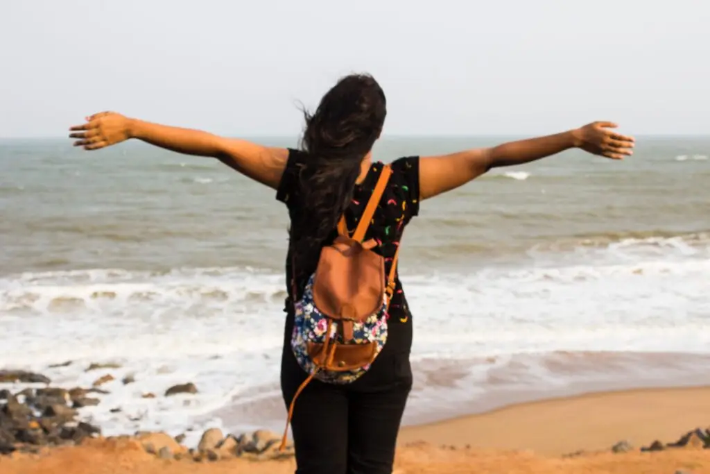 Woman with floral backpack standing on beach with arms outstretched, facing the ocean waves.