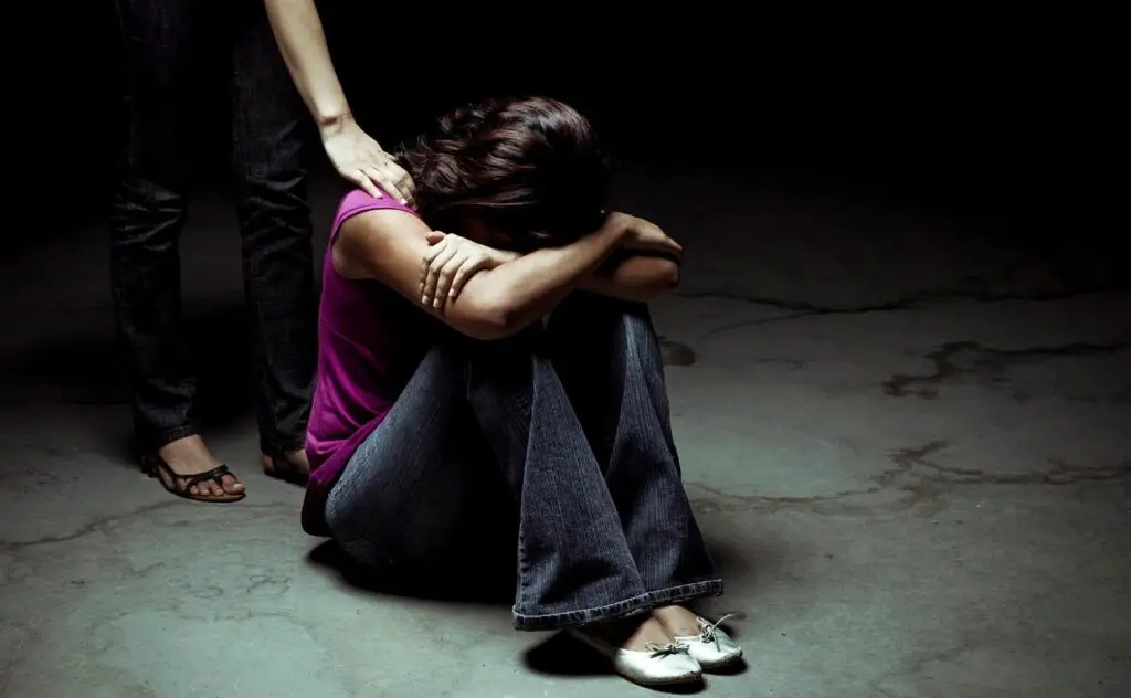 Distressed woman sitting on the floor with head down while another person offers a comforting hand on her shoulder.