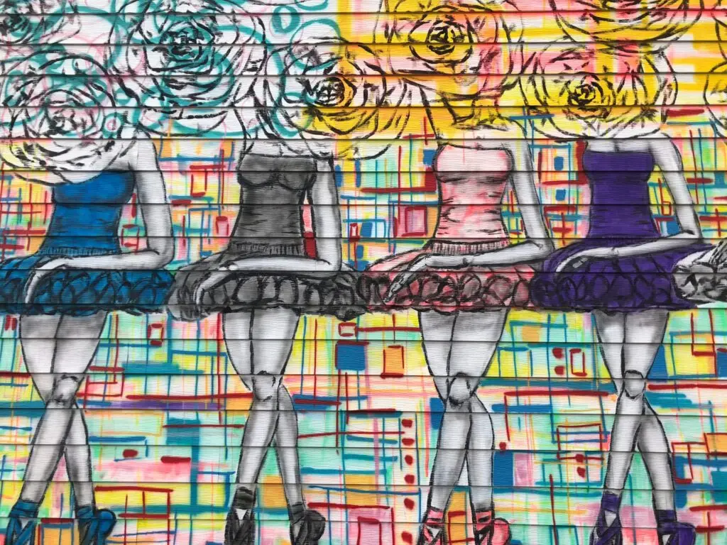Colorful mural of four ballerinas in tutus against an abstract floral and grid-patterned background.