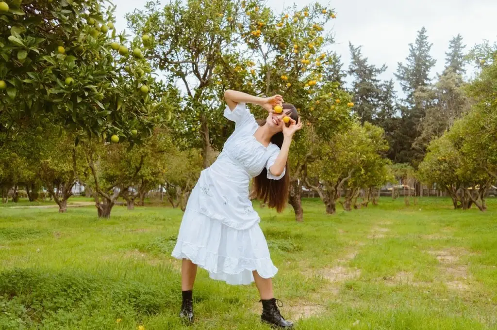 Woman in white dress holding oranges over her eyes in a citrus orchard with green grass.